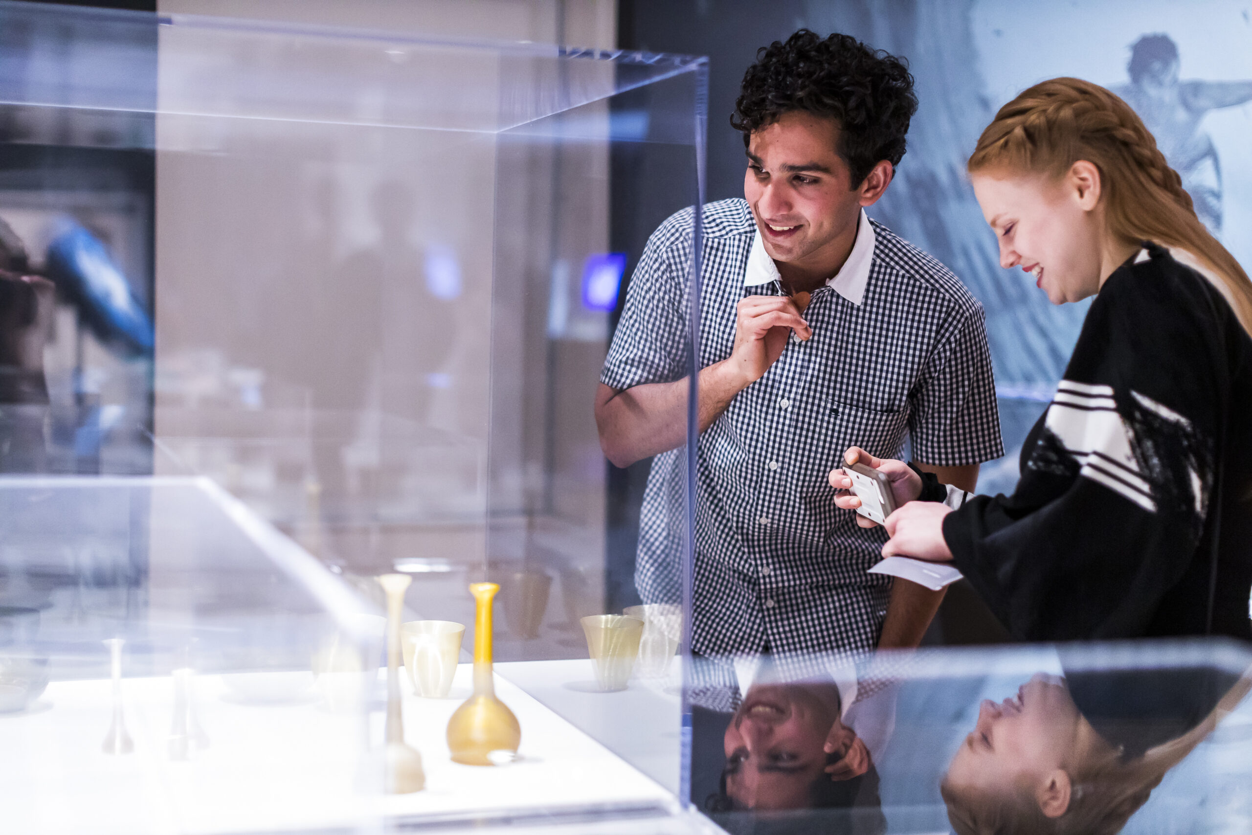 Two visitors look at 3D printed vases behind a glass case. One visitor smiles as she takes a photo with her phone.