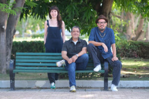 A group of three people sitting outdoors on and around a green bench, with trees and bushes in the background.