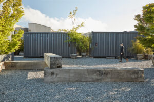 A gravel lot with large bench-like pieces of timber lined by potted trees and a set of large gray shipping containers.