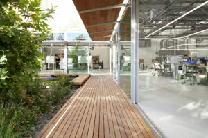 A courtyard garden lined with wooden benches, a shaded slanted overhang, and walking path surrounded by a factory interior with floor-to-ceiling windows that double as sliding doors.