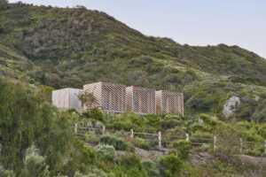 Four large cubic open-air structures made with slanted wood paneling sit nestled in the green foliage of a rocky canyon hillside.