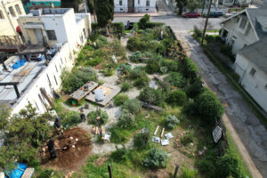 Overhead view of garden lot under construction with people digging a mound of dirt, scattered building and landscaping debris, a pair of people standing in the vegetation, and another pair unloading a bin from a truck parked outside the garden's entrance.