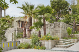 An exterior view of a home constructed from concrete blocks with a series of layered terraces lined with palm trees, cacti, and blooming vegetation.