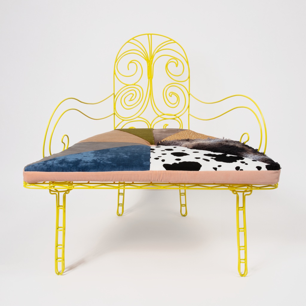 A chair composed of yellow-painted metal for its back, arms, and legs, with the back and arms bent into round, whimsical shapes. The seat is upholstered with multiple fabrics quilted together.